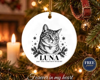 May include: A round white ceramic ornament featuring a black line drawing of a cat. The text reads LUNA, 2010-2026, and FOREVER IN MY HEART. The design includes small snowflakes and pine branches, suspended by a gold cord.