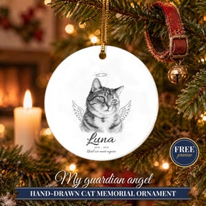 May include: A round white ceramic ornament featuring a black and white sketch of a cat with angel wings and a halo. The text reads Luna, 2010-2026, and Until we meet again. The ornament hangs from a gold cord and is designed for display.
