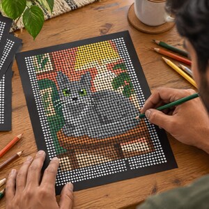May include: A dot-to-dot coloring sheet featuring a gray cat sitting on a surface, rendered in a grid of numbered circles. The image includes a yellow lamp, green foliage, and a brown table, with several colored pencils scattered nearby.
