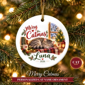 May include: A round white ceramic Christmas ornament featuring a sleeping tabby cat on a plaid blanket. The text reads Merry Catmas, Luna, and 2026. The illustration includes a window, a small decorated tree, a lantern, and a mug.