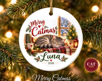 May include: A round white ceramic Christmas ornament featuring a sleeping tabby cat on a plaid blanket. The text reads Merry Catmas, Luna, and 2026. The illustration includes a window, a small decorated tree, a lantern, and a mug.
