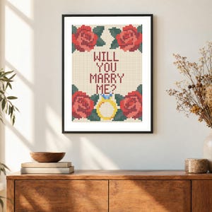 May include: A framed cross-stitch embroidery featuring the text WILL YOU MARRY ME? in dark red letters, surrounded by four red roses with green leaves and a yellow engagement ring with a blue gemstone, all set against a cream-colored grid.