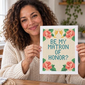 May include: A rectangular white card featuring a cross-stitch pattern with the text BE MY MATRON OF HONOR? in blue letters. The design includes pink roses with green leaves in the four corners on a light beige grid background.