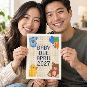 May include: A rectangular white sign featuring a pixelated cross-stitch design with the text BABY DUE APRIL 2027 in dark blue. The design includes a blue baby onesie, a cluster of colorful balloons, a yellow rubber duck, and a brown teddy bear.