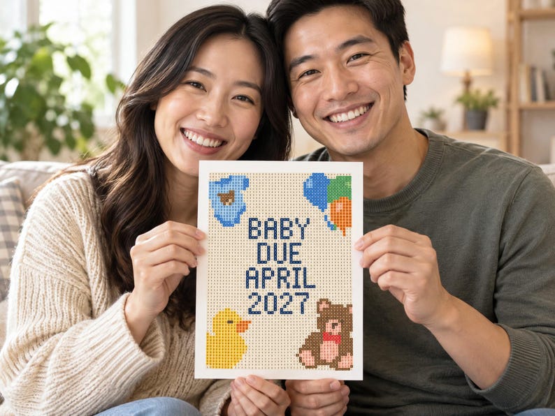 May include: A rectangular white sign featuring a pixelated cross-stitch design with the text BABY DUE APRIL 2027 in dark blue. The design includes a blue baby onesie, a cluster of colorful balloons, a yellow rubber duck, and a brown teddy bear.