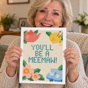 May include: A rectangular print featuring a pixelated design with the text YOU'LL BE A MEEMAW! in blue. The design includes a red teapot, a yellow lemon, pink flowers with green leaves, and a blue teacup on a light beige grid background.