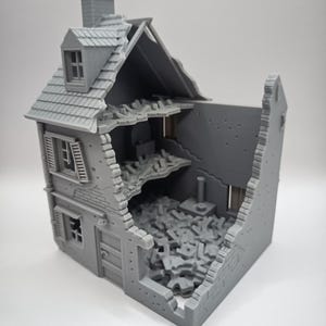 May include: A grey model of a two-storey house, showing significant damage. The roof is partially collapsed, and the walls are broken, exposing the interior. The model includes a chimney, windows, and a door.