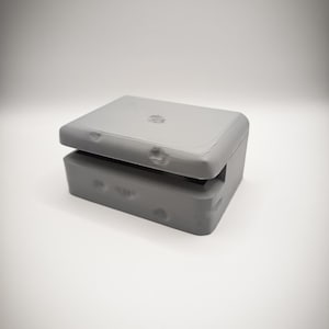 Peut inclure: Une boîte rectangulaire grise avec un couvercle à charnière, légèrement entrouvert. La boîte a une finition lisse et mate et semble être faite d'un matériau solide. Le design est simple et moderne, avec des bords arrondis et un petit élément décoratif sur le couvercle.