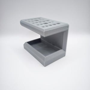 Peut inclure: Un organisateur gris imprimé en 3D avec de multiples trous circulaires sur le dessus et un plateau rectangulaire en dessous. L'organisateur est conçu pour contenir divers articles, tels que des fournitures d'art ou des outils. Le design est moderne et minimaliste.