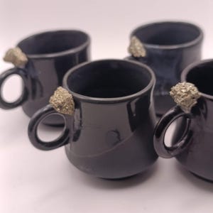 May include: Four black ceramic mugs with a glossy finish. Each mug features a unique, textured, gold-colored accent on the handle. The mugs are arranged on a white surface, showcasing their dark color and distinctive design.