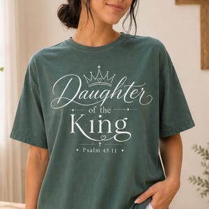 Könnte beinhalten: Ein dunkelgrünes Kurzarm-T-Shirt mit weißem Aufdruck Daughter of the King, einer Kronengrafik über dem Wort Daughter und einem kleinen Kreuzsymbol unter dem Wort King, gefolgt von dem Text Psalm 45 11.