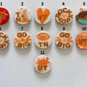 May include: Eleven circular pins featuring orange and white designs with gold glitter accents. The pins display text including UT, TN, ROCKY TOP, VOLS, GO BIG, and UTK, alongside graphics of stars, a heart, a football, and a mountain landscape.