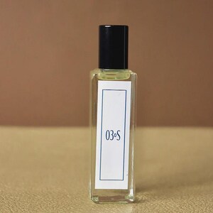 Puede incluir: Un frasco de perfume rectangular de vidrio con tapa negra. El frasco tiene una etiqueta blanca con borde azul y el texto "03.S". El frasco está lleno de un líquido amarillo claro.