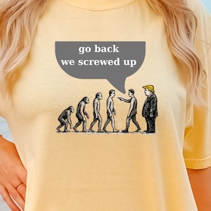 Puede incluir: Camiseta amarilla con un gráfico que representa la evolución humana, culminando en una figura que se asemeja a un político. Una burbuja de diálogo sobre las figuras dice "go back we screwed up".