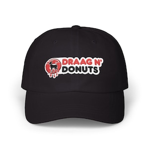 Op de afbeelding: Een zwarte baseballpet met een geborduurde patch op de voorkant. De patch toont een rode donutafbeelding met een zwart silhouet van een hert en de tekst "DRAAG N' DONUTS" in dikke witte en zwarte letters met een druipend rood effect.