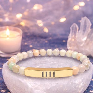 May include: A beaded bracelet with a gold-colored bar featuring the number "1111". The bracelet is made of light-colored, round beads. The background includes a lit candle and soft, blurred lights.