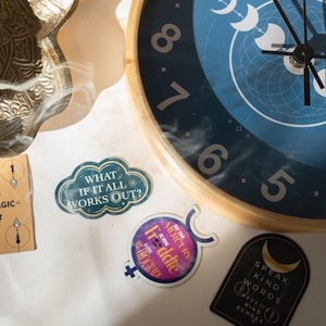 May include: A close-up of a blue and wooden clock with white numbers and moon phase designs. Several stickers are scattered around the clock, including one that says "WHAT IF IT ALL WORKS OUT?" and another that says "Freddie".