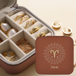 May include: A brown jewelry travel case with a gold Aries symbol and the name "Sarah" printed on the front. The open case reveals various gold rings, earrings, and other jewelry pieces. The case is pink and has a gray interior.