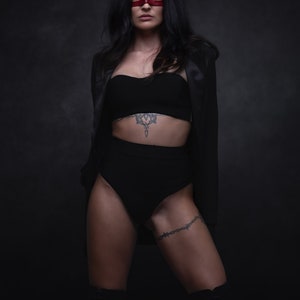 Puede incluir: Una mujer con una chaqueta de sat&eacute;n negro, sujetador negro y pantalones cortos negros de cintura alta. Lleva una m&aacute;scara roja sobre los ojos y tiene un tatuaje en el est&oacute;mago y el muslo. Est&aacute; de pie frente a un fondo oscuro.