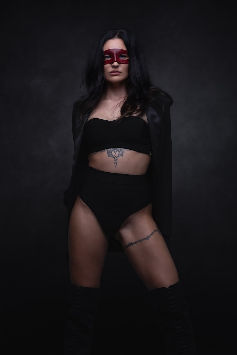 Puede incluir: Una mujer con una chaqueta de sat&eacute;n negro, sujetador negro y pantalones cortos negros de cintura alta. Lleva una m&aacute;scara roja sobre los ojos y tiene un tatuaje en el est&oacute;mago y el muslo. Est&aacute; de pie frente a un fondo oscuro.