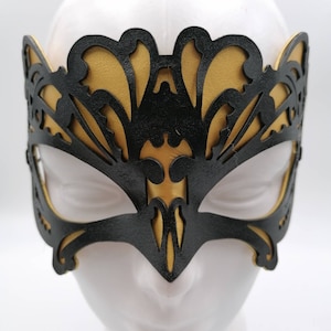 May include: A black and gold masquerade mask with an intricate, cut-out design. The mask features a black overlay with a gold backing, creating a striking contrast. The mask is designed to cover the eyes and forehead, with a pointed lower edge.