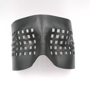 以下が含まれることがあります： 一連の正方形の切り抜きが施された、濃いグレーの革製アイマスク。マスクは顔にフィットするように湾曲しており、上部は幅広く、下部は狭くなっています。素材は滑らかで、わずかに反射しているように見えます。