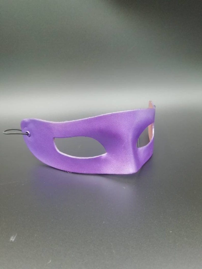 Puede incluir: M&aacute;scara de carnaval morada con un solo agujero para el ojo. La m&aacute;scara est&aacute; hecha de un material liso y brillante y tiene un dise&ntilde;o simple y elegante.