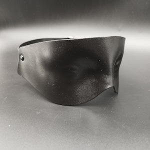 Puede incluir: Máscara para los ojos de cuero negro con forma curva y un solo botón en el lateral.