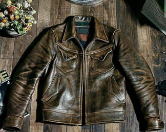 May include: A brown leather jacket with a distressed finish. The jacket features a collared neckline, multiple pockets, and buttoned cuffs. The jacket is laid flat on a wooden surface.