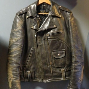 May include: A distressed black leather motorcycle jacket featuring a wide notched collar, silver-tone metal snap buttons, and multiple zippered pockets. The jacket has a quilted interior lining, a front zip closure, and visible wear on the sleeves.