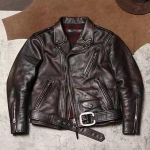 Peut inclure: Veste en cuir marron foncé de style motard classique. Elle présente un col cranté, une fermeture éclair asymétrique, plusieurs poches zippées et une ceinture à boucle argentée. La veste a un aspect usé et vieilli.