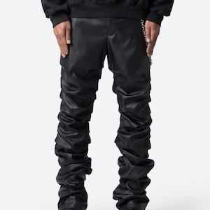 Puede incluir: Pantalones de cuero negros con diseño fruncido y detalle de cadena. Los pantalones tienen cintura alta y se combinan con botas negras. El estilo general es atrevido y moderno.