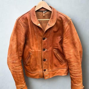 May include: An orange-brown leather jacket with a classic collar and button-up front. The jacket has two front pockets and buttoned cuffs. It hangs on a wooden hanger against a grey background. The jacket appears vintage.