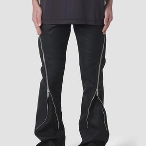 Puede incluir: Pantalones negros con cremalleras plateadas que recorren las costuras exteriores. Las cremalleras se extienden desde el tobillo hasta la cintura. Los pantalones tienen un ligero ensanchamiento en la parte inferior y se llevan con zapatos negros.