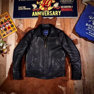 Puede incluir: Chaqueta de cuero negra con cremallera, un pequeño bolsillo en el pecho y dos bolsillos laterales. La chaqueta se muestra sobre una superficie marrón con un fondo de madera. Una pancarta arriba dice "40 ANNIVERSARY".