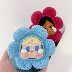 Puede incluir: Dos muñecas hechas a mano con forma de flor de ganchillo. Una muñeca está con un disfraz de flor azul claro con pelo rubio y ojos azules. La otra muñeca está con un disfraz de flor roja con pelo negro y una cara llorando. Ambas muñecas están sobre una superficie plateada.