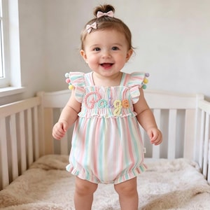 May include: A baby wearing a pastel striped romper with the name "Paige" embroidered on the chest. The romper has ruffled sleeves and leg openings, and is adorned with colorful pom-poms. The baby is smiling and wearing a pink bow in their hair.