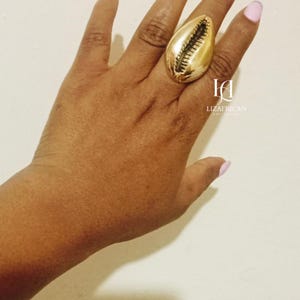 May include: A large, gold-colored ring in the shape of a seashell is worn on a hand with light pink nail polish. The ring is the focal point, with the brand name "LIZAFRICAN" visible in the background.