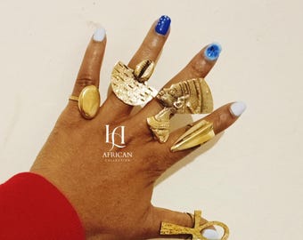 May include: A hand displaying several gold-toned rings, including an oval signet ring, a ring with a seashell, a ring with a bust, and a ring with an ankh symbol. The nails are painted in shades of blue. The text "AFRICAN COLLECTION" is visible.