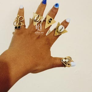 Op de afbeelding: Een hand die een verzameling goudkleurige ringen toont. De ringen hebben verschillende ontwerpen, waaronder geometrische vormen, spiralen en versieringen. Sommige ringen bevatten zwarte en witte accenten en schelpen. De tekst "African Collection" is zichtbaar.