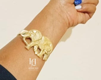 May include: Gold-colored elephant cuff bracelet. The bracelet features a detailed elephant design and is worn on a wrist. The image also shows the text "African Collection".