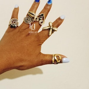 May include: A hand adorned with multiple gold rings. The rings showcase diverse designs, such as spirals, a seashell, and a cross. The image also includes the text "AFRICAN COLLECTION".
