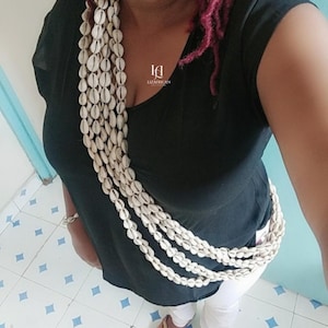 Puede incluir: Un collar de varias hebras hecho de conchas blancas. El collar se lleva sobre una camiseta negra. La imagen también muestra el texto "LIZAFRICAN COLLECTION".