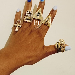 May include: A hand displaying several gold-colored rings with various designs, including an ankh, a spiral, a triangle, and a seashell. The nails are painted in shades of blue. The image includes the text "AFRICAN COLLECTION".