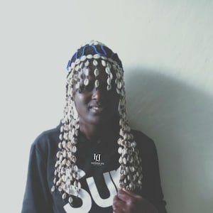 Può includere: Un copricapo nero e blu con perline e conchiglie bianche pendenti. La persona indossa una felpa nera con la scritta "SU" a blocchi bianchi. Il logo "LIZAFRICAN" è stampato sulla felpa.