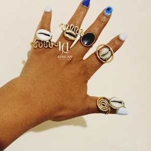 Puede incluir: Una mano que muestra varios anillos dorados con diseños únicos. Los anillos presentan conchas de cauri, espirales y formas abstractas. La imagen incluye el texto "AFRICAN COLLECTION".
