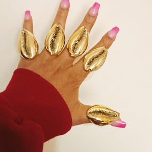 May include: A hand wearing five large, gold-colored shell rings. The nails are painted with a pink ombre design. The sleeve of a red sweater is visible in the image.