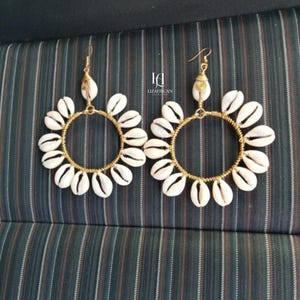 May include: A pair of gold-toned hoop earrings adorned with white seashells. The earrings feature a circular design with the shells arranged around the hoops. The earrings are displayed against a striped background.