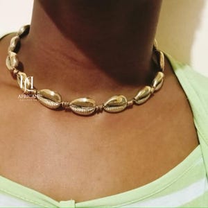 May include: A gold-colored necklace made of connected shell-shaped beads. The necklace is worn on a person wearing a light green shirt. The necklace is a statement piece and is likely handmade.