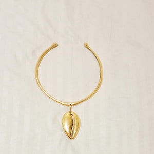 Pode incluir: Um colar gargantilha dourado com um pendente em forma de concha. O colar tem uma banda circular que se abre na frente. O pendente de concha é liso e brilhante, com uma textura detalhada na frente.